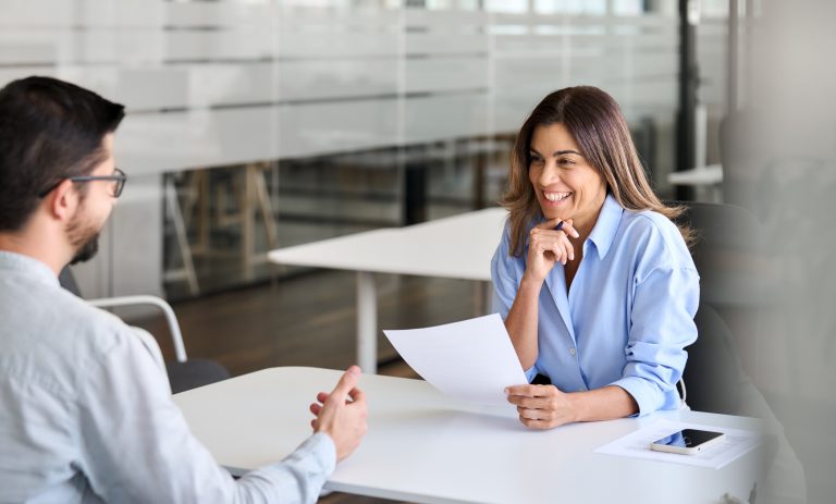 Apartment shopping, Apartment Mystery Shops, Apartment renters Journey A woman in a blue shirt sits at a table with a man, holding papers and smiling. They appear to be having a meeting in a modern office environment. Multifamily Journey Lens CX for the Apartment Industry