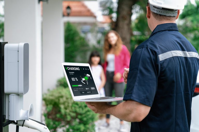 Multifamily Maintenance Management TechnologyApartment renters Journey A technician adept in maintenance management holds a laptop showing a 60% battery charging status near an electric vehicle charging station, with two people in the background. Multifamily Journey Lens CX for the Apartment Industry