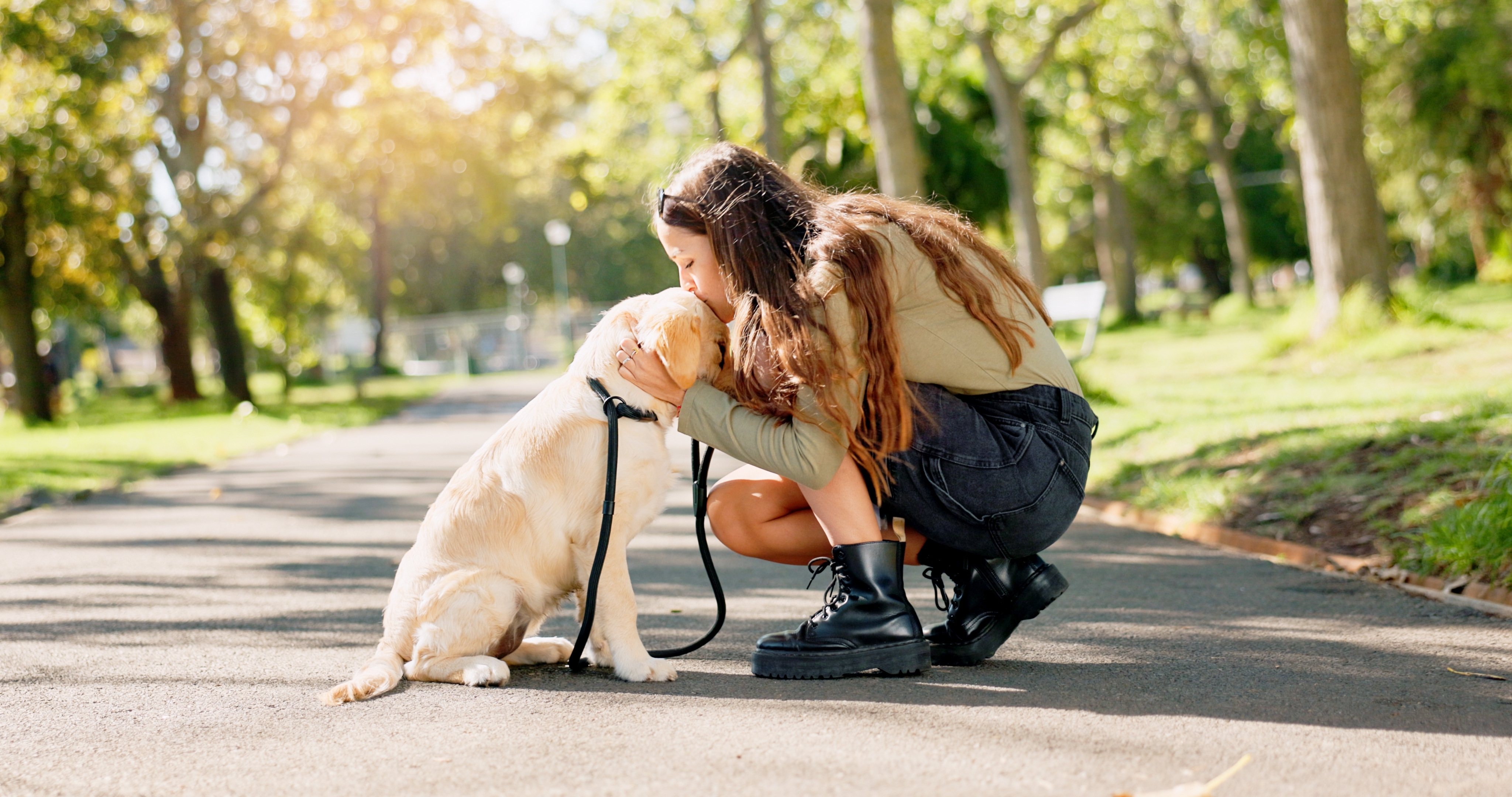 Apartment shopping, Apartment Mystery Shops, Apartment renters Journey A woman kneels on a park path, lovingly holding a seated Labrador wearing a leash. Trees and sunlight fill the background. Multifamily Journey Lens CX for the Apartment Industry