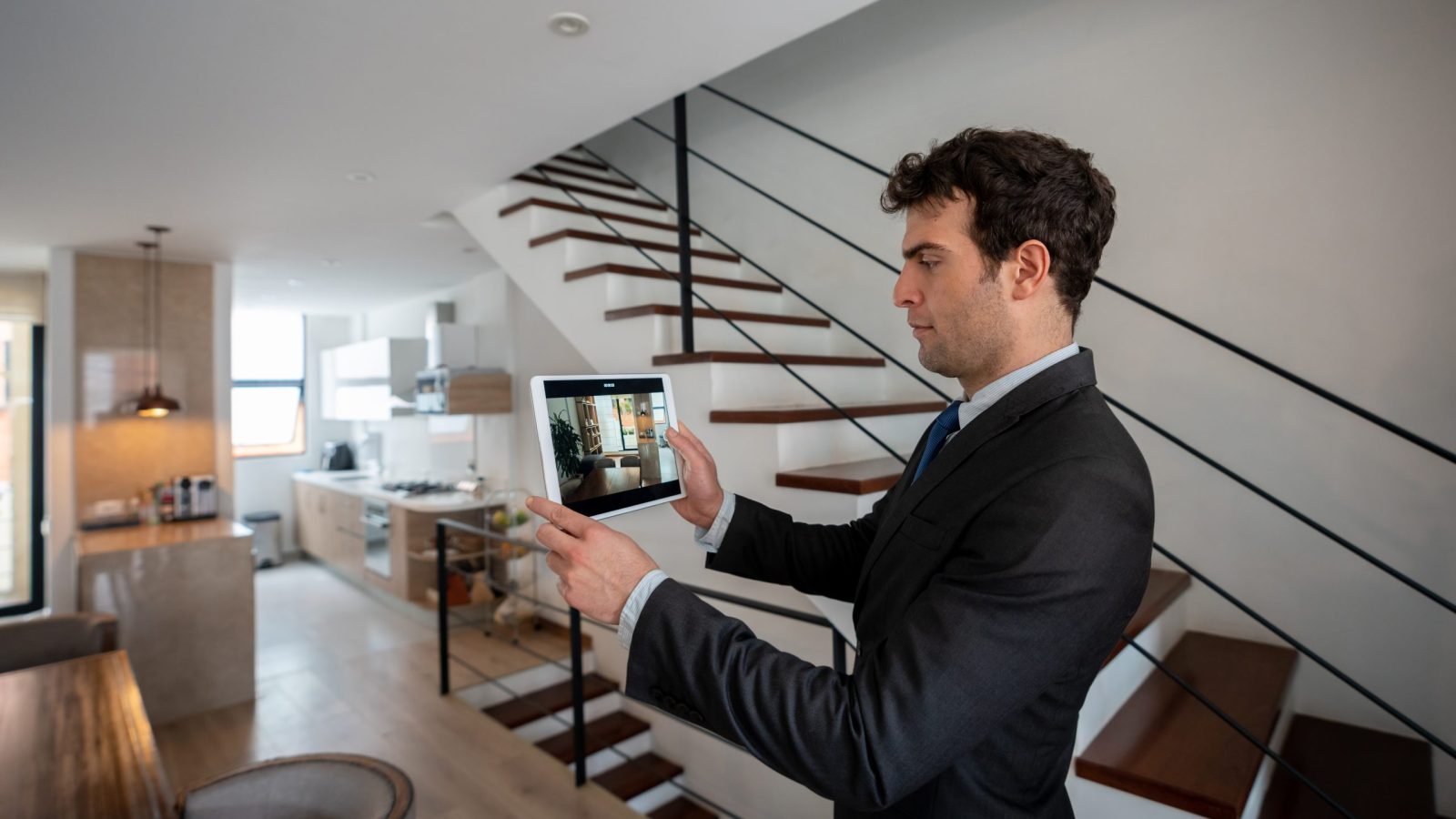 Apartment shopping, Apartment Mystery Shops, Apartment renters Journey A man in a suit captures user feedback on a tablet while standing in a modern home interior, featuring a sleek staircase and open-plan kitchen. Multifamily Journey Lens CX for the Apartment Industry